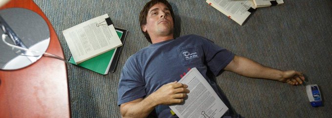 A young man in a blue t-shirt lying on a carpeted floor surrounded by scattered books, papers, and notebooks.