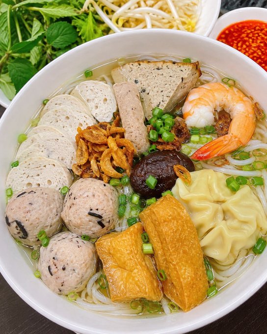 A bowl of Vietnamese-style noodle soup topped with shrimp, meatballs, wontons, tofu, fried shallots, and fresh herbs, served with bean sprouts, mint, and chili sauce on the side.