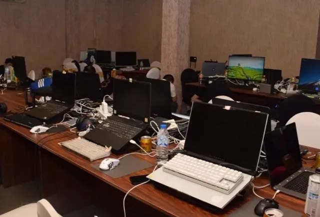 A dimly lit cyber café or co-working space is filled with rows of laptops and desktop computers, with several people working at the stations amid tangled cables, water bottles, and keyboards scattered across long wooden tables. The image loosely symbolizes the digital operations behind the Asia Express news story about large-scale crypto processing and emerging crypto-backed financial products in Asia.