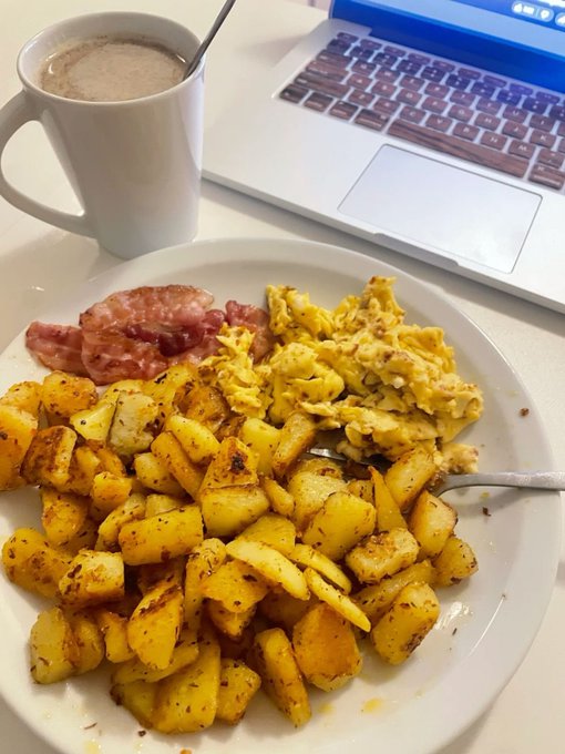 A breakfast plate with scrambled eggs, bacon, and seasoned potatoes sits next to a coffee mug and a laptop keyboard.