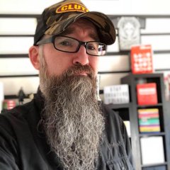 A bearded man wearing glasses and a cap stands in what appears to be a store.