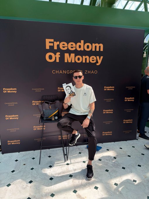 A man sits in front of a "Freedom Of Money" banner referencing Changpeng Zhao (CZ), holding his book, wearing an Adidas t-shirt and sunglasses. The image relates to the post about BNB meme season, as CZ is the former CEO of Binance — the company behind the BNB Chain ecosystem where memecoins are actively traded.