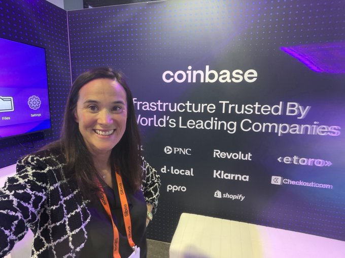 A woman with dark hair wearing a black and white tweed jacket and an orange lanyard smiles in front of a Coinbase booth displaying 'Infrastructure Trusted By World's Leading Companies' along with logos for PNC, Revolut, eToro, Klarna, Shopify, and others.