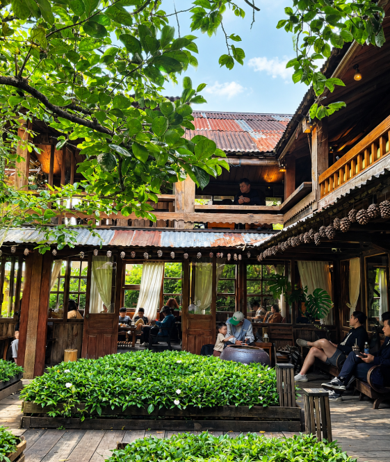 A rustic wooden cafe or restaurant with an open courtyard, surrounded by lush green plants and trees, with patrons seated at tables and on a balcony.