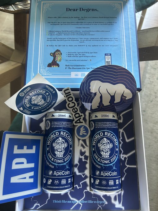 The image shows a box containing two cans of beer, one blue and one white, with stickers on them that say "World Record" and "Apecoin". The box also has a note attached to it that says "Dear Degenics", which is likely a message from someone who sent the gift.
