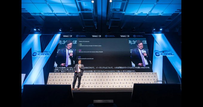 A man in a suit stands on a stage giving a presentation at a Web3 event, with large screens behind him showing 'CRYL' branding and Japanese text '光3C' along with a 'Table of Contents' slide.