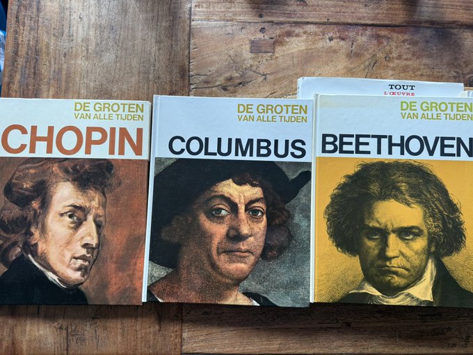 Three books titled 'DE GROTEN VAN ALLE TIJDEN' featuring portraits of Chopin, Columbus, and Beethoven lie on a wooden table.