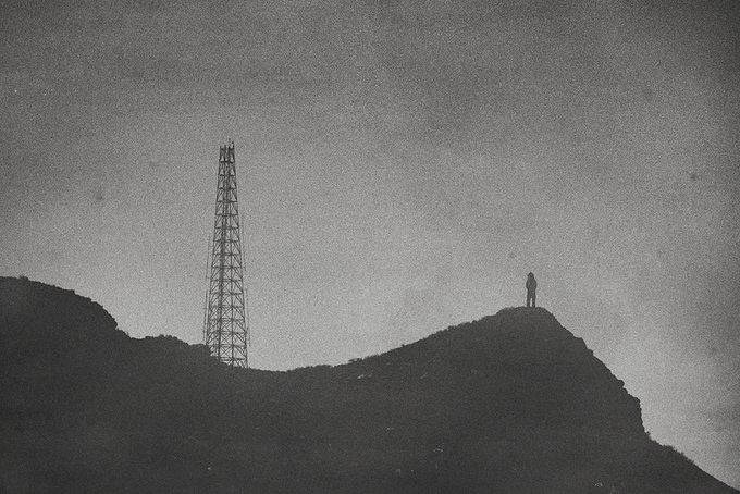 A grainy, black-and-white image of a lone figure standing on a rocky hill next to a transmission tower under a cloudy sky.