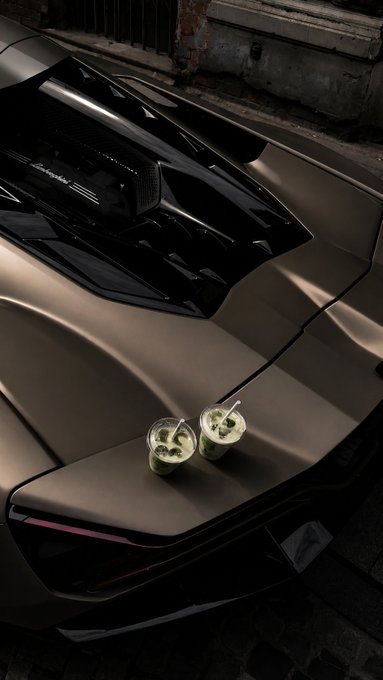 Two green matcha drinks with straws sit on the rear engine cover of a champagne-colored Lamborghini sports car parked on a city street.