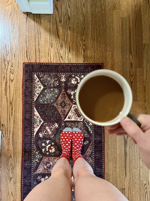 A first-person view of someone wearing red and white heart-patterned socks standing on a decorative rug, holding a mug of coffee with cream, with wooden floors visible.