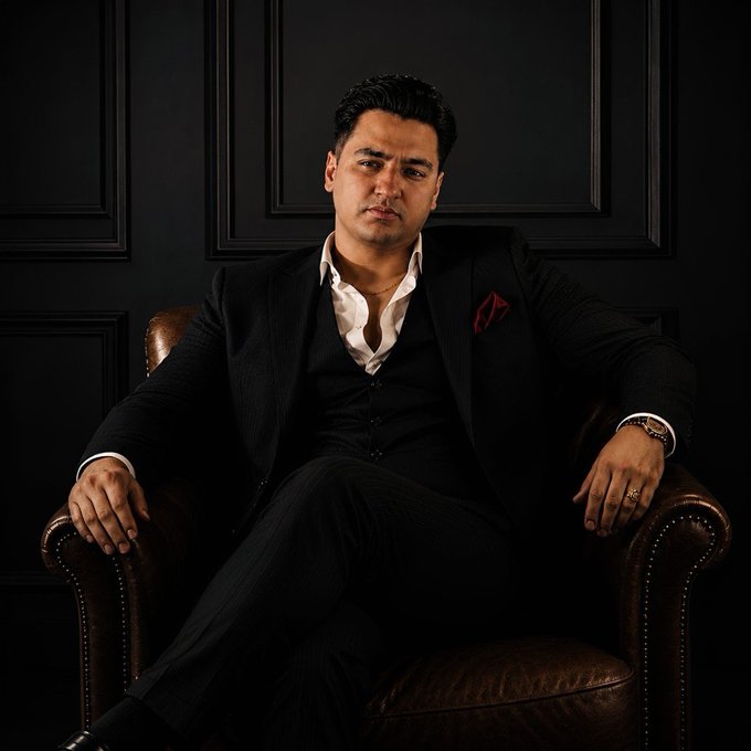 The image features a man sitting in a chair, dressed in a suit with a red pocket square. The man has his arms crossed over his chest and appears to be looking off into the distance or deep in thought. The background of the photo is black, which contrasts with the white shirt he's wearing. There are no other people visible in the image, and it seems like the focus is on the man sitting alone in the chair.