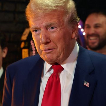 President Donald Trump, wearing a navy suit with a red tie and an American flag lapel pin, looks on with a serious expression while a smiling man stands behind him. The image accompanies a post quoting his remarks about the $TRUMP memecoin, in which he acknowledges hearing it performs well while distancing himself from running it.
