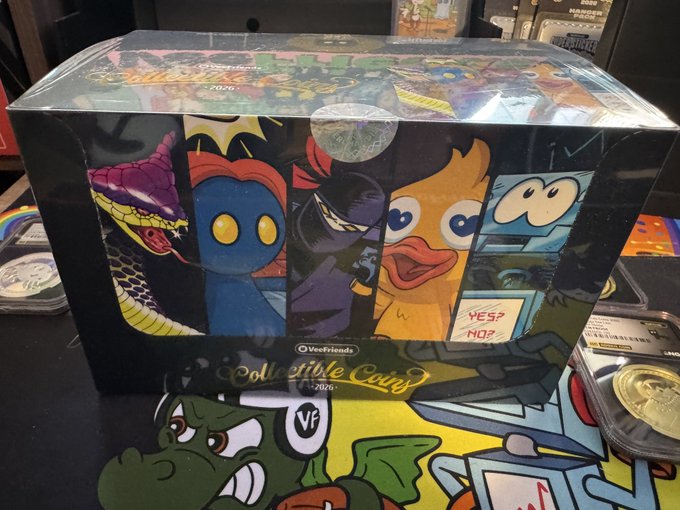 A sealed display box of VeeFriends 'Collectible Coins' featuring character artwork on the packaging, sitting on a table.