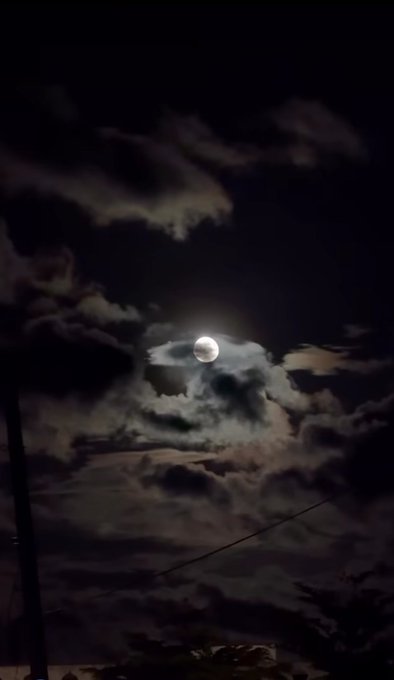 A bright full moon shines through thick, dramatic clouds in a dark night sky, with a utility pole and power lines visible in the foreground.