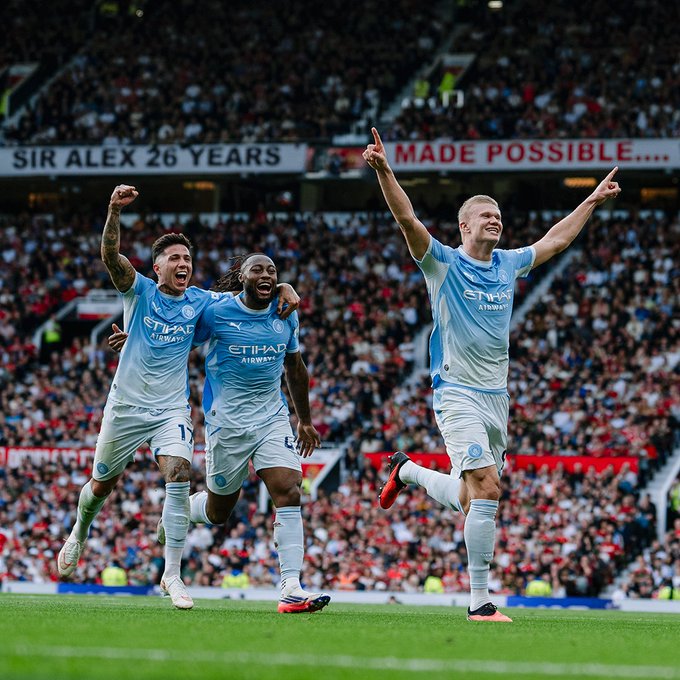 Three Manchester City soccer players in light blue Etihad Airways jerseys celebrate on the pitch with their arms raised, in front of a stadium crowd holding a banner reading 'SIR ALEX 26 YEARS MADE POSSIBLE...'.
