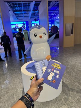 A hand holding a 'FOOD FACTORY' promotional card with a penguin design, in front of a penguin mascot statue at what appears to be an event or exhibition.