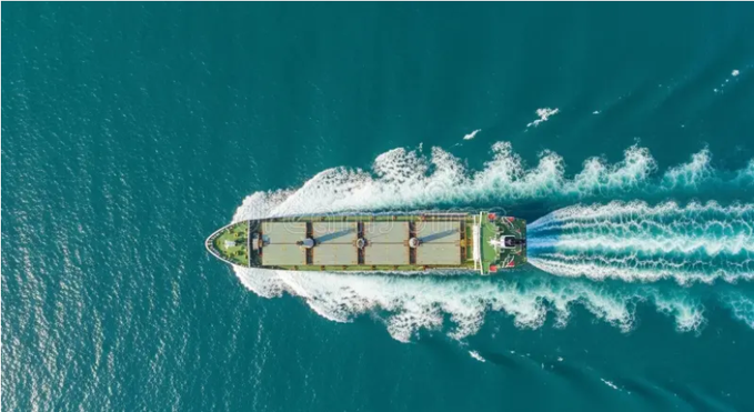 An aerial view of a large cargo ship sailing through deep teal ocean water, leaving a foamy white wake behind it.