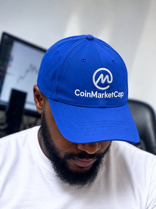 A man wearing a blue CoinMarketCap branded baseball cap looks downward, with trading charts visible on monitors in the background.
