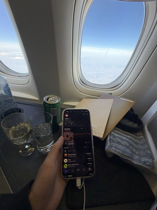 A first-person view from an airplane window seat showing a hand holding a smartphone displaying a crypto portfolio app (Coin Stats) with a value of $9,229, alongside a glass of amber liquid, a water glass, a green soda can, and a notebook.