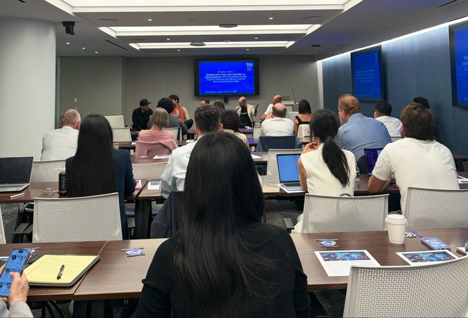 A conference room scene with attendees seated at desks facing two screens displaying presentation slides, with laptops and coffee cups visible on the tables.