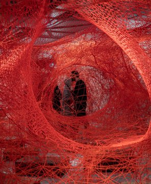 Two silhouetted figures wearing hats stand within a circular opening surrounded by intricate, swirling red threads against a dark background.