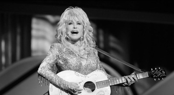 Black and white photo of Dolly Parton performing on stage with an acoustic guitar, wearing a sparkling sequined outfit.
