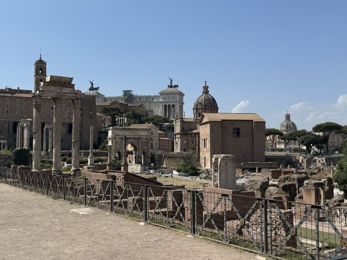A scenic view of ancient Roman ruins featuring columns, the Arch of Septimius Severus, and domed buildings under a clear blue sky.