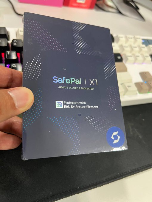 The image shows a person holding up a new item, possibly a computer mouse or keyboard, with a sticker on its back that says "safe pal 1". The person's hand is visible above the item, while the background features a desk setup with a keyboard and a mouse. There are also other keyboards in the scene, suggesting that this might be an office environment where multiple people work together.