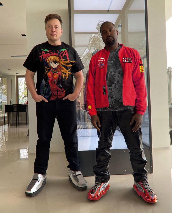 Two men stand indoors; the left wears an Evangelion Asuka t-shirt and silver shoes, while the right wears a red jacket and red Yeezy Foam Runner sneakers.
