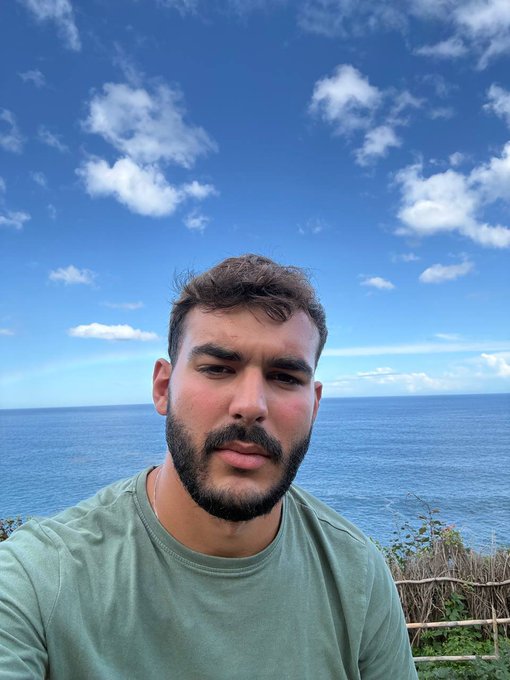 A bearded man in a green t-shirt takes a selfie against a stunning backdrop of blue ocean and scattered clouds, embodying a relaxed "good morning" vibe that pairs perfectly with the optimistic "GM crypto fam" greeting.
