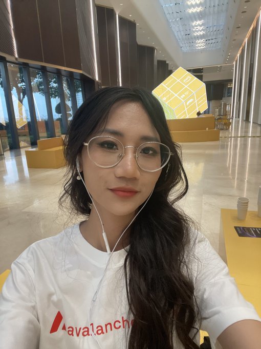 A young woman wearing glasses and white earphones takes a selfie while wearing a white t-shirt with the red 'Avalanche' logo, standing in a modern building lobby.