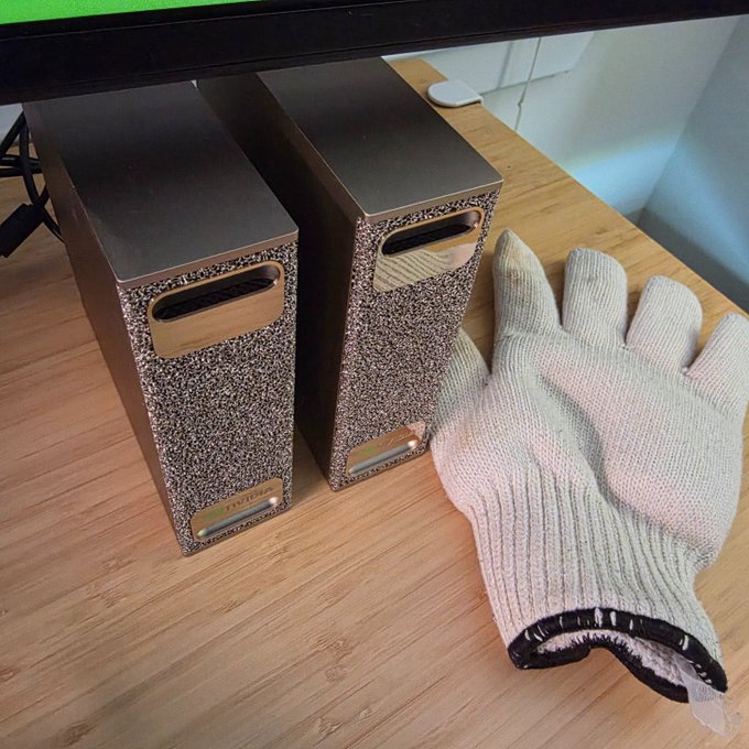 A pair of silver metallic speakers with textured speckled fronts sit on a wooden desk next to a gray knit work glove, placed there to safely handle the speakers which became extremely hot after 8 hours of running a quantization workload.
