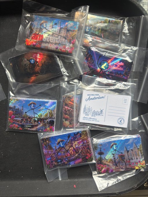 The image shows a collection of nine small, colorful postcards scattered across a black surface. Each card features a unique design or illustration, ranging from cityscapes with buildings and bridges to various cartoon characters. The postcards are neatly arranged on top of each other in two rows of four, creating an interesting visual effect that draws attention to the vibrant designs. The text "postcard" is clearly visible, indicating the purpose of these items as souvenirs or mementos from a special event or gathering.