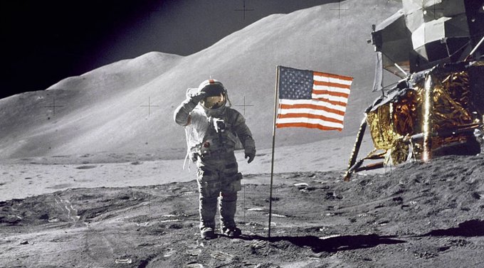 An astronaut in a white spacesuit saluting beside an American flag on the surface of the moon, with a lunar lander module visible to the right.