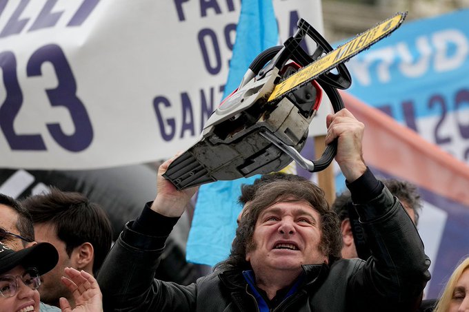 The image shows a man holding up a chainsaw with a banner above his head that says "It's great that this debate in @ethereum is finally happening in the open. The agenda of @trent_vanepps is to get funding to keep the current Ethereum core dev teams working". A crowd of people surrounds him, and they are all holding up signs with messages related to the ongoing debate on Ethereum.