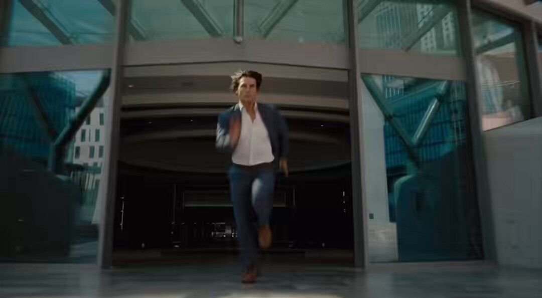 The image shows a man running through an open glass door, possibly entering or leaving a building. He appears to be dressed in a suit and tie, suggesting that he might be a businessman or attending a formal event. The scene takes place indoors with large windows surrounding him.