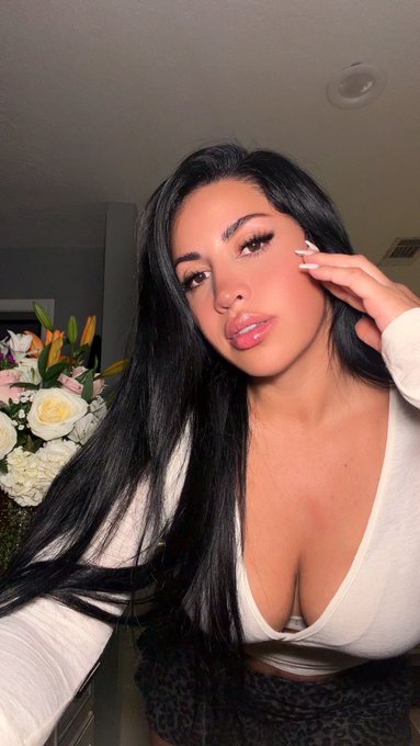 A selfie of a woman with long dark hair wearing a white low-cut top, posing indoors with a vase of flowers visible in the background.
