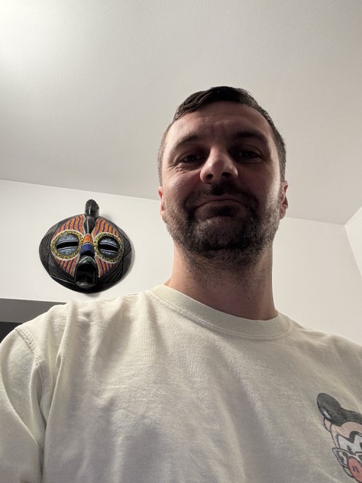 A selfie of a bearded man in a white t-shirt with an African-style decorative mask mounted on the wall behind him.
