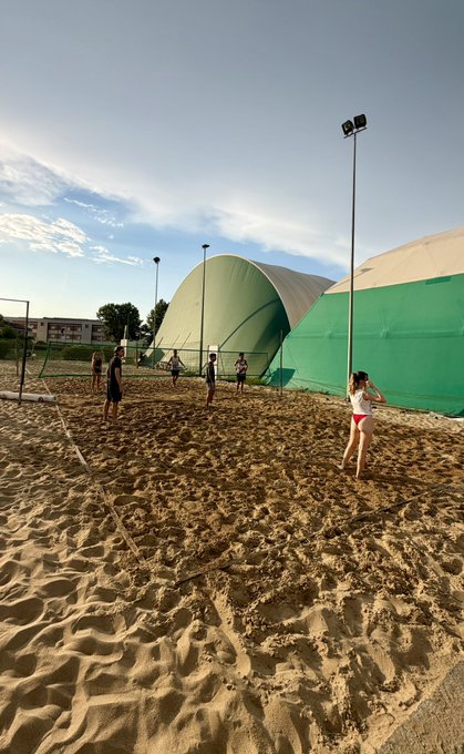 A casual beach volleyball game takes place on a sandy court with green domed sports facilities in the background under a partly cloudy sky.