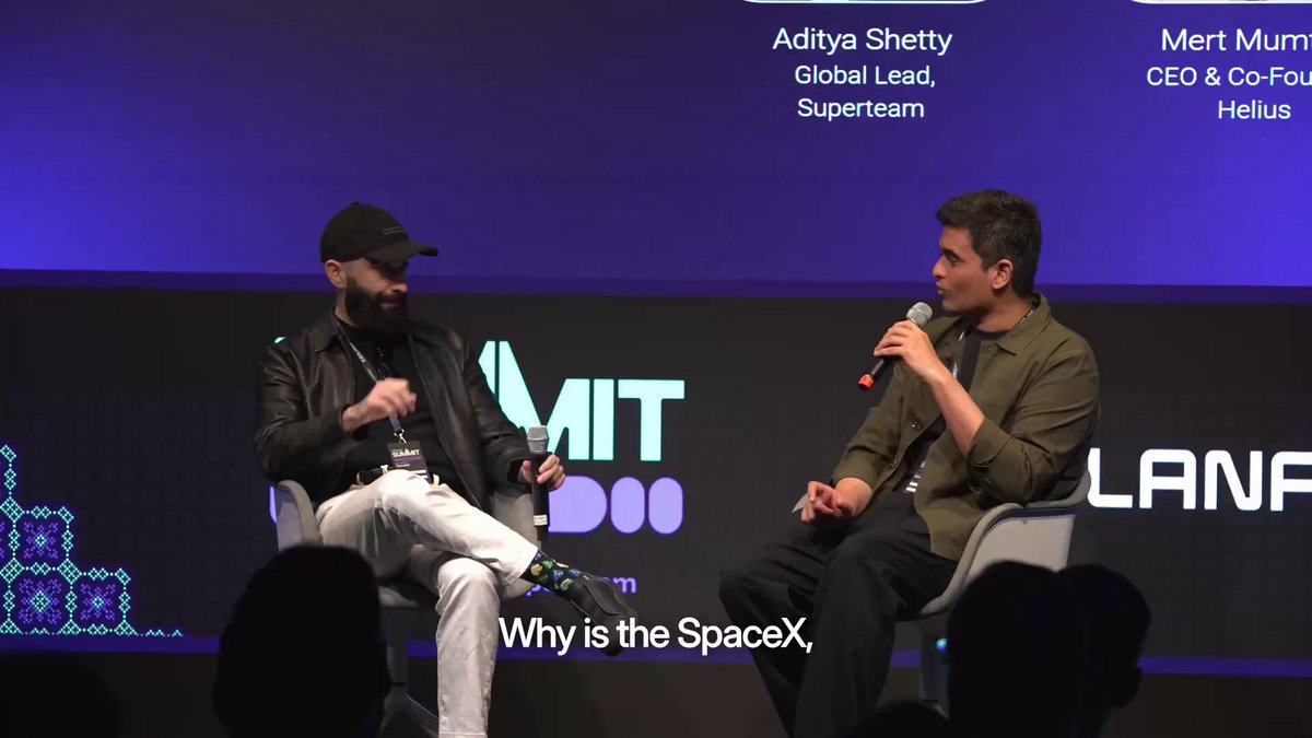 The image shows two men sitting next to each other, talking to a camera. They are wearing hats and appear to be discussing something related to space exploration or technology. The background features a large screen displaying the words "Why is the SpaceX?" written in white text on a blue background. This suggests that they might be discussing the latest developments in space exploration or the impact of new technologies, such as the Tesla-powered Mars Rover.