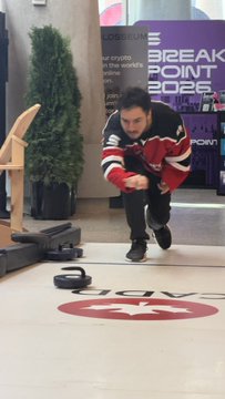 A man in a red and white hockey jersey prepares to throw a curling stone down a rink with a 'BREAKPOINT 2026' banner visible in the background.