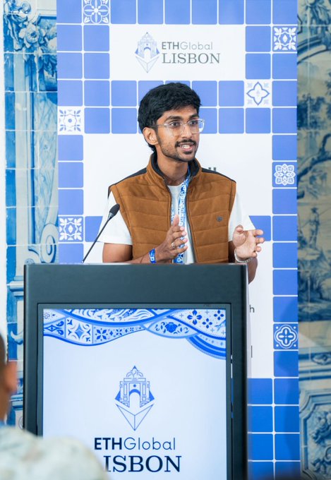 A young man wearing glasses and a brown vest speaks at a podium during the ETHGlobal Lisbon event, set against a blue tiled backdrop.
