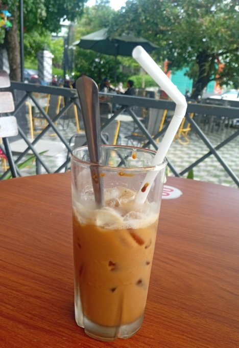 A nearly empty glass of iced Thai milk tea with a spoon and a bent white straw, sitting on a wooden table at an outdoor cafe with tables and umbrellas visible in the background.