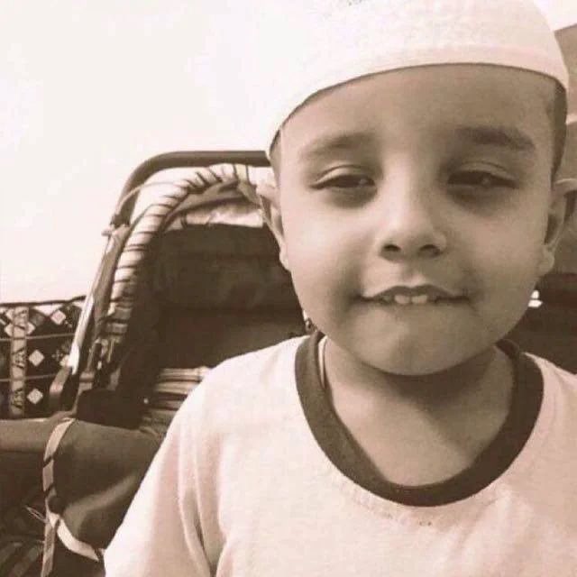 A sepia-toned photograph of a smiling young child wearing a white cap and shirt, sitting on what appears to be a patterned chair.