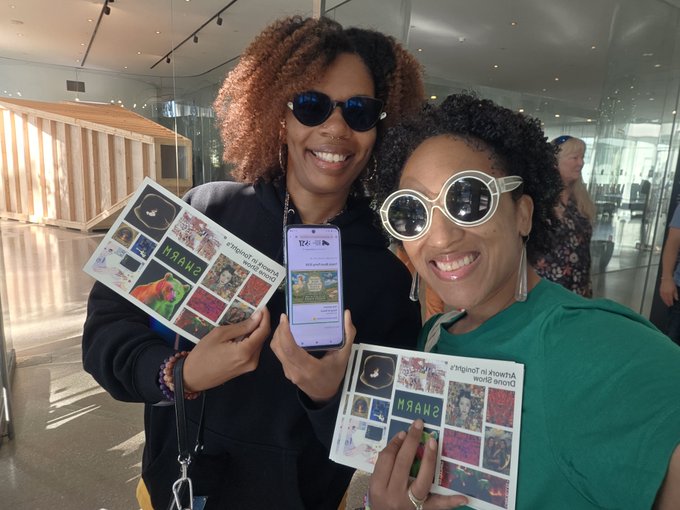 Two women at an event smiling and holding promotional materials that say 'SHAHI Always in Favor of Drama' and display a phone screen.