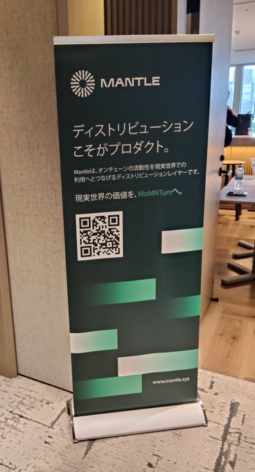 The image features a large green and white banner that says "AI Meets Crypto" written in Japanese, with an image of a computer screen behind it. The banner also includes a QR code for a website called "Mantle Tokyo Meetup". In the background, there is a person sitting at a desk, which could be part of a meeting or event related to AI and cryptocurrency.