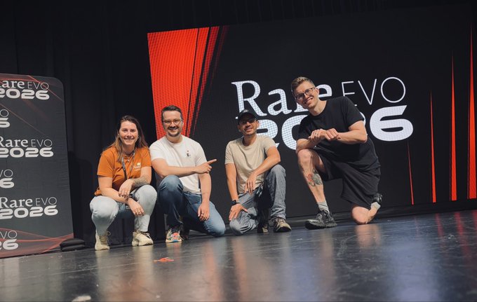 Four people posing together on stage at the Rare EVO 2026 conference, with three kneeling/crouching and one jumping, set against a backdrop displaying the event's red and white logo.