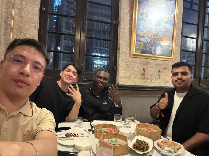 Four men sit together at a restaurant table filled with dim sum dishes, posing for a photo with drinks and bamboo steamers scattered across the table.