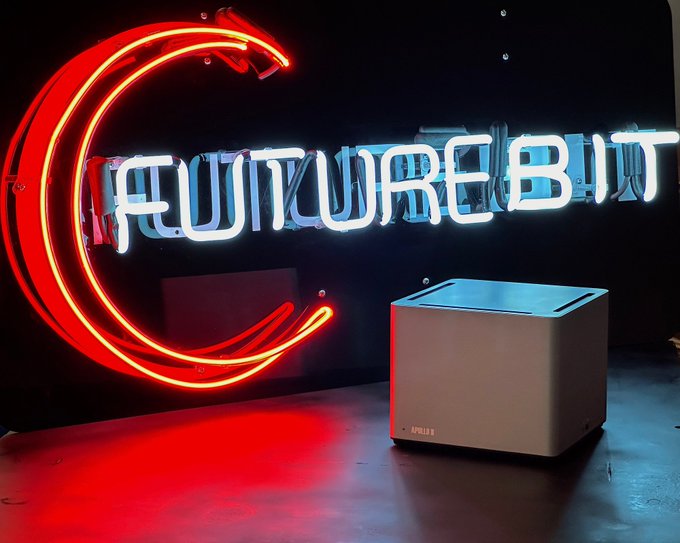 A neon sign displaying the text "FUTUREBIT" with a red crescent moon design, positioned next to a small white mining machine on the floor.