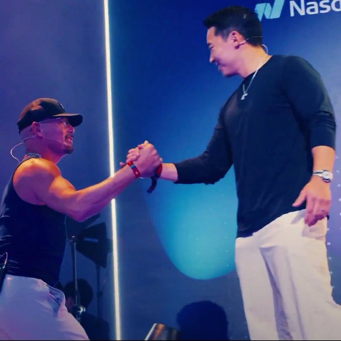 Two men clasp hands on a brightly lit stage; a partial “Nasdaq” logo is visible in the background.